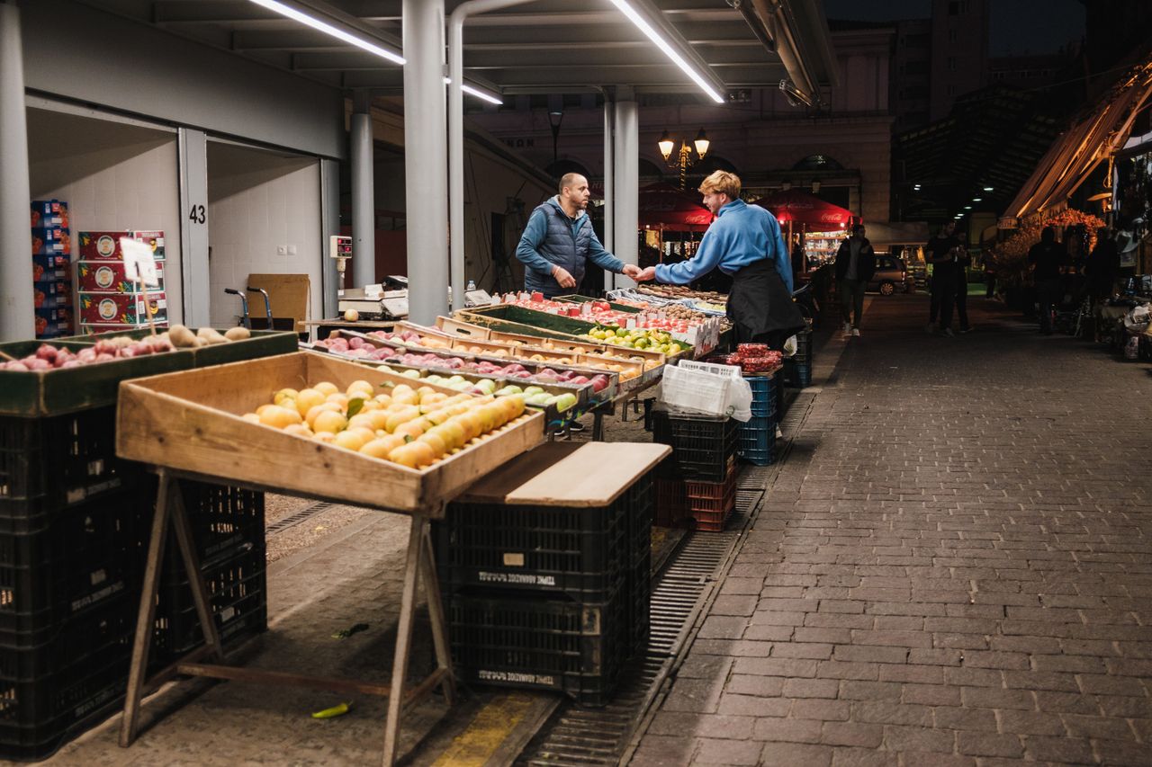 Two people exchanging goods at a well-lit night market in the city, surrounded by fresh produce.